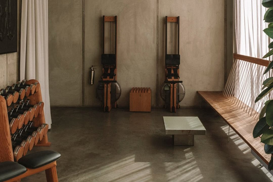 The private studio — concrete floor, wooden weight rack, rowing machines, linen curtains, a wooden bench by the window.